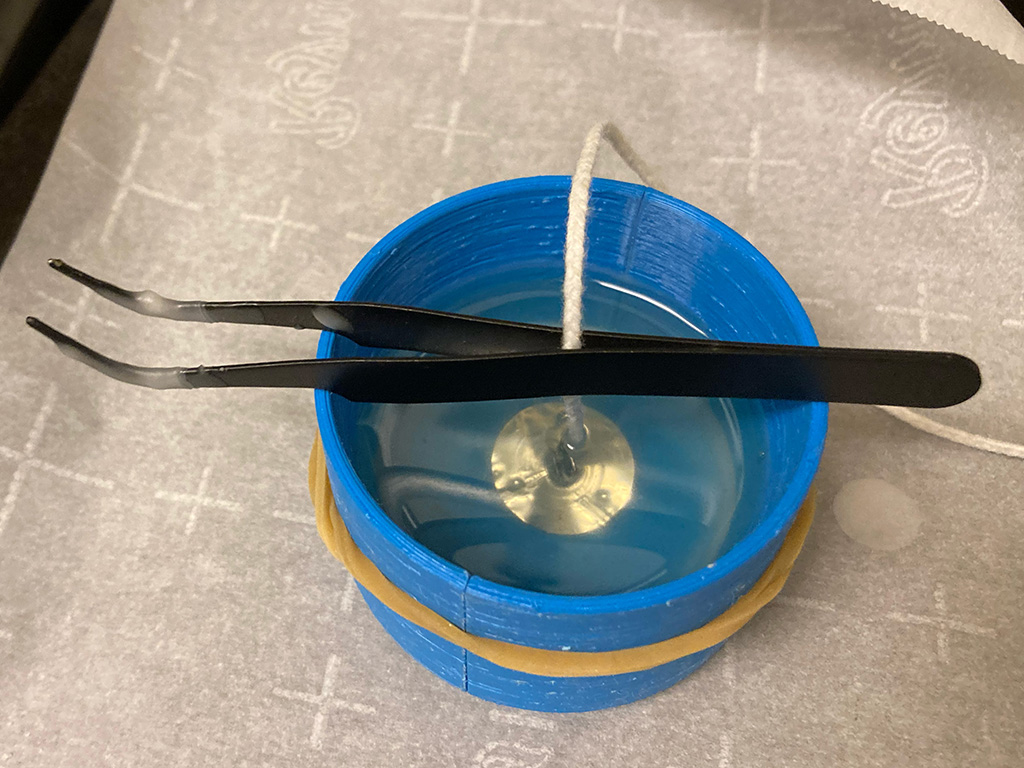 Photo of mold containing translucent wax; a pair of tweezers suspends a wick and metal base in the middle of the molten wax