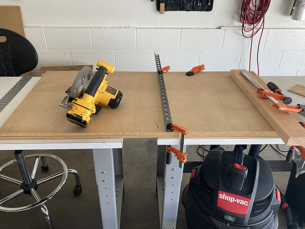 Photo of particle board laying across two workbenches with a gap to allow the circular saw to cut the board; a metal rail is clamped to the board to guide the saw