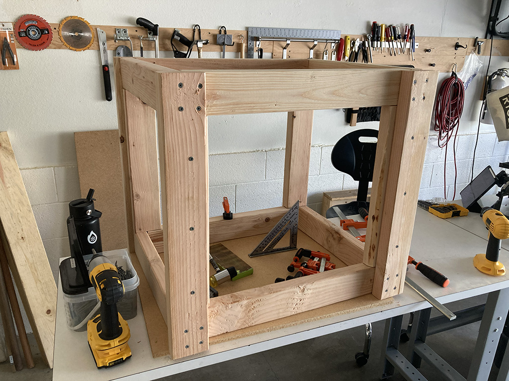 Photo of assembled box without top or bottom on top of workbench; many tools are around and sitting within the box