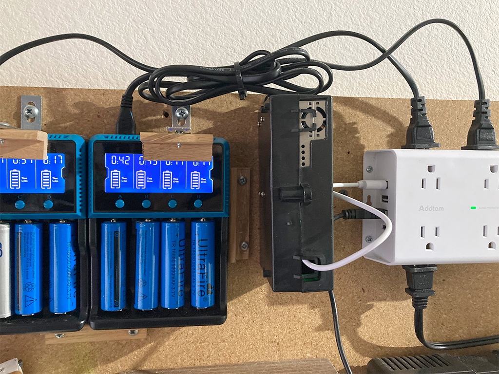 Assembled case mounted on charging wall with a short USB cable plugged into a power strip; two battery chargers are visible on the left.