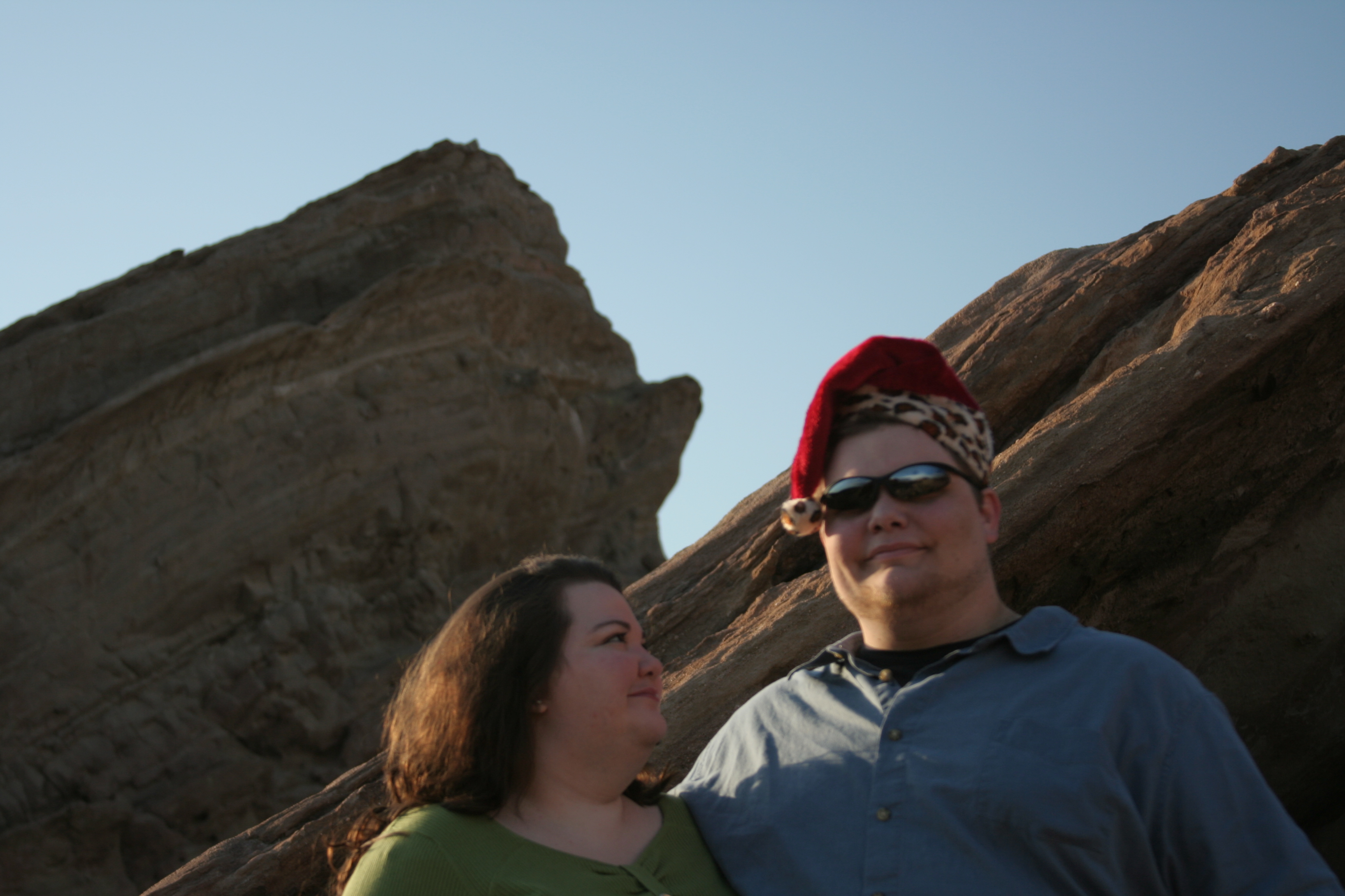 Bonnie and Daniel from 2009 at Vasquez Rocks. Bonnie is looking intently at Daniel, and Daniel is wearing a leopard print fuzzy Santa cap and sunglasses.